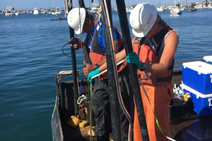 Two people in white hard hats on a boat handling core sampling equipment